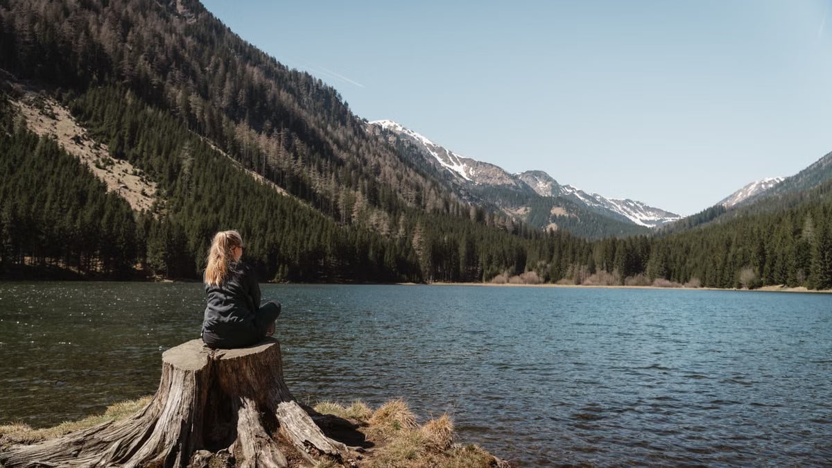 A young woman sits on a tree stump by a tranquil lake surrounded by snow-capped mountains.